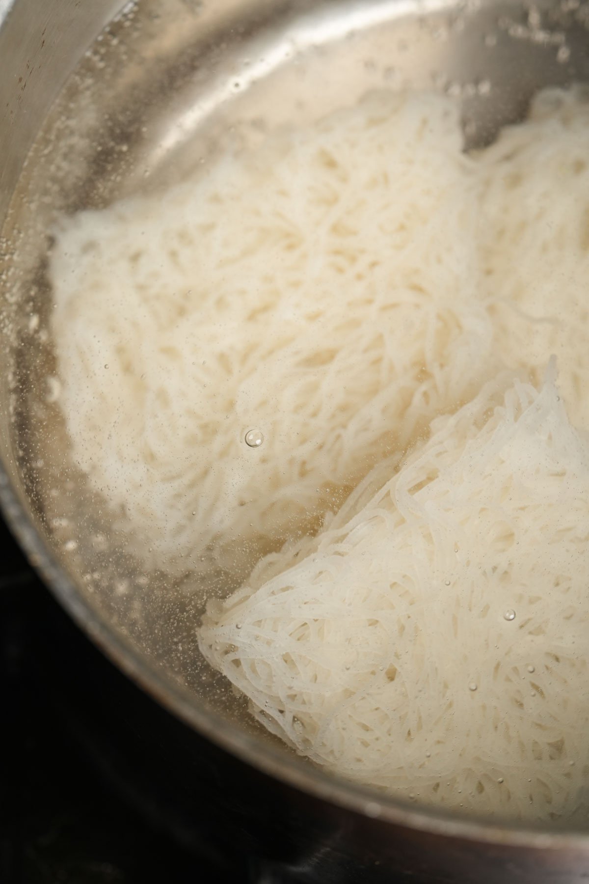Rice noodles boiling in a pot of water, with bubbles forming on the surface as the noodles cook.