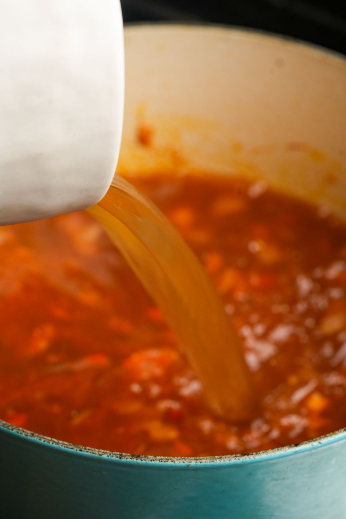A close-up of vegan bun rieu broth being poured from a white pitcher into a pot of red soup with visible vegetables, inside a teal-colored pot.