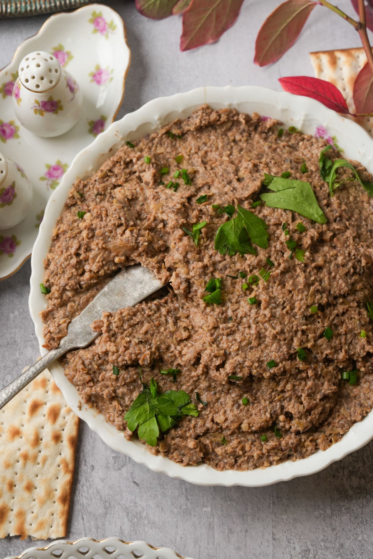 A white bowl filled with a chunky brown vegan chopped liver garnished with fresh parsley. A vintage butter knife rests inside the pate. Crackers and floral-patterned salt and pepper shakers are placed nearby on a gray surface.