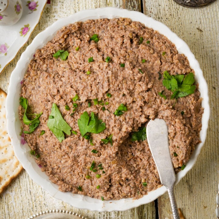 A bowl of vegan chopped liver spread garnished with parsley, with a butter knife resting on the side, on a rustic white wooden table next to pieces of matzo.