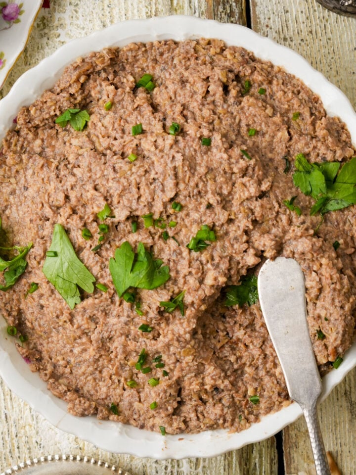A bowl of vegan chopped liver spread garnished with parsley, with a butter knife resting on the side, on a rustic white wooden table next to pieces of matzo.