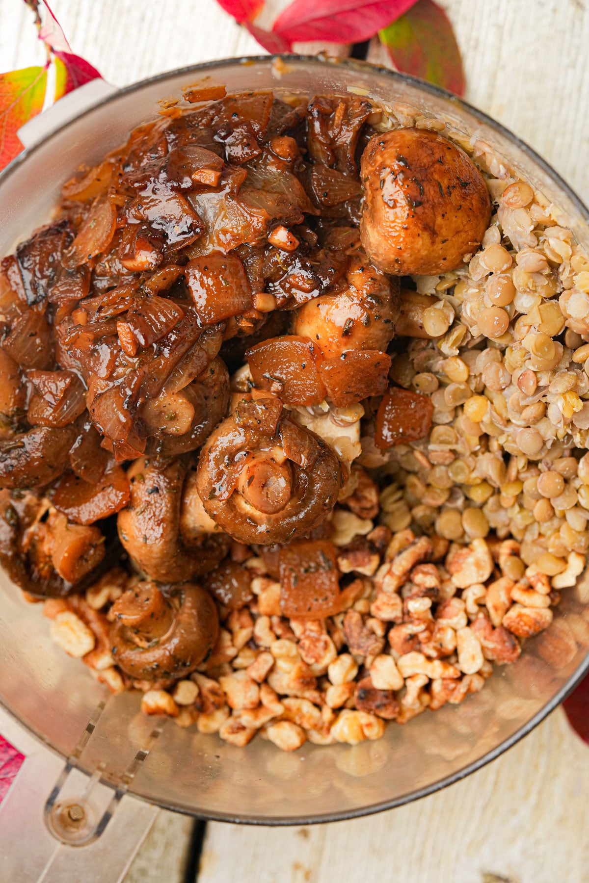A food processor bowl filled with cooked brown lentils, sautéed mushrooms and onions, walnuts, and seasonings, ready to be blended. The bowl is placed on a light wooden surface with red autumn leaves nearby.