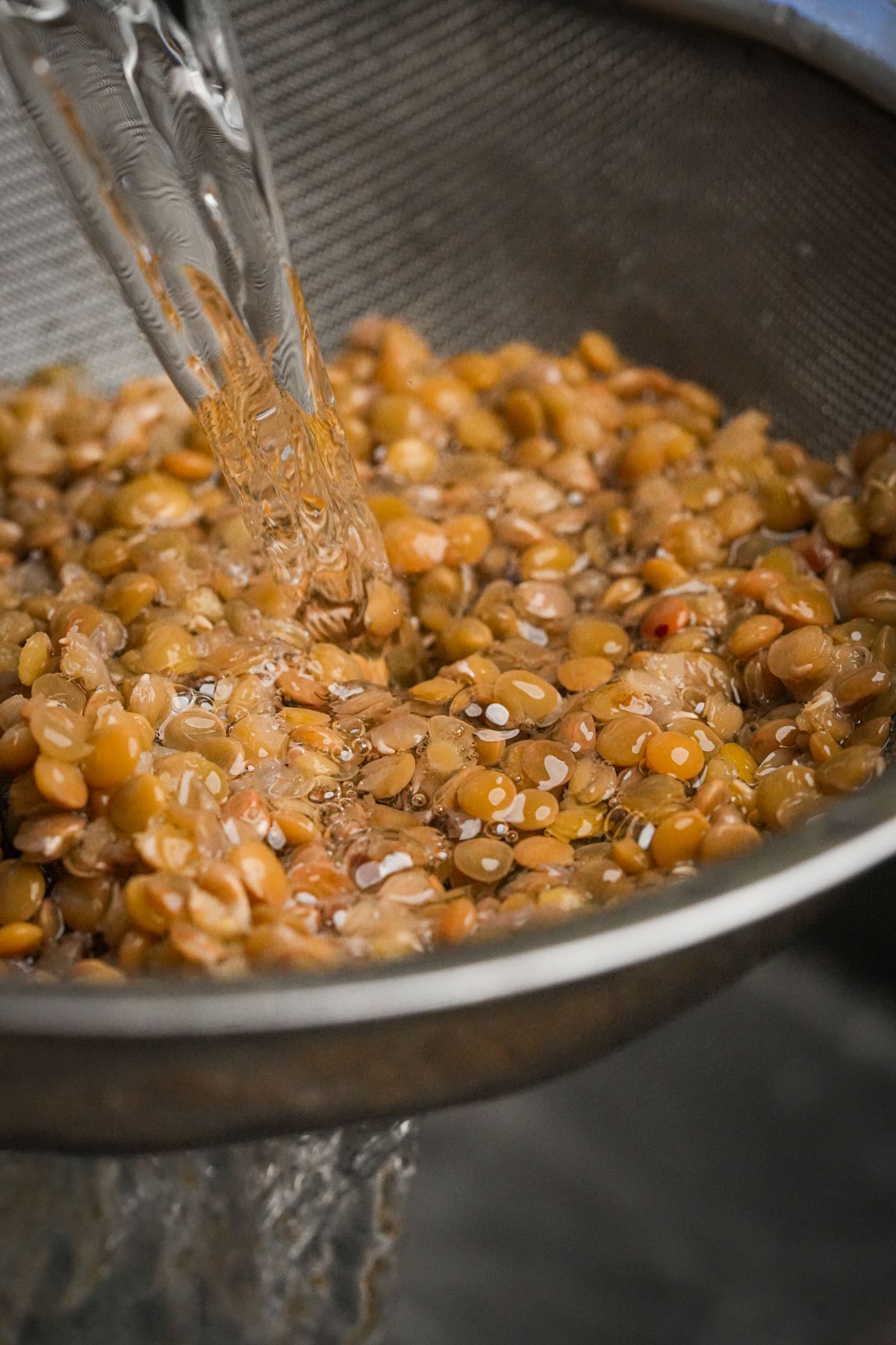 Water is being poured over brown lentils in a metal mesh strainer, rinsing them thoroughly.