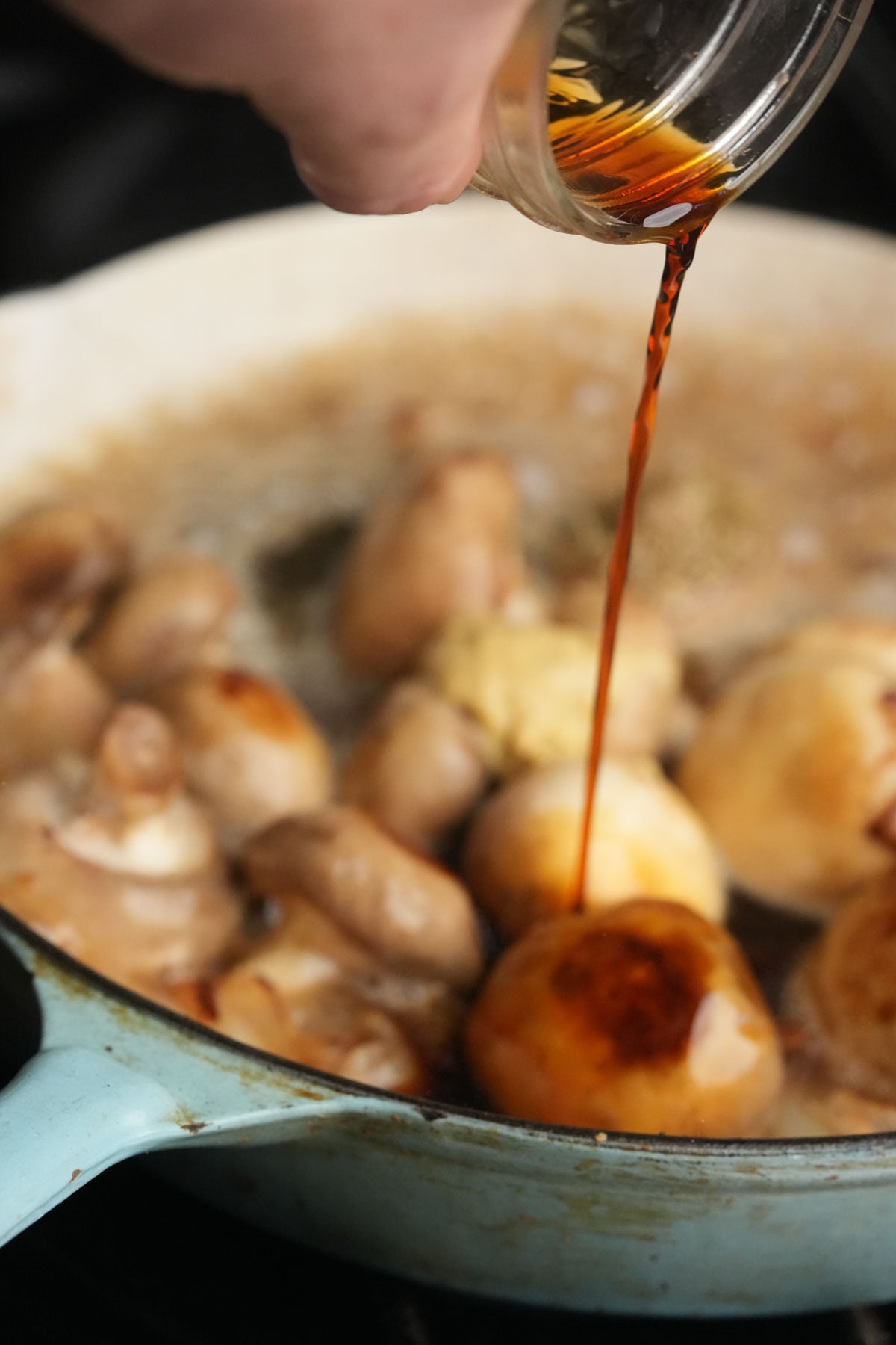 A close-up of a hand pouring a tamari from a glass jar over whole mushrooms and onions cooking in a light blue pan. Steam rises from the sizzling ingredients.