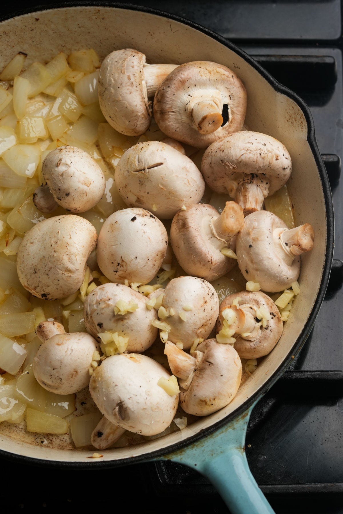 Whole mushrooms, chopped onions, and minced garlic are being sautéed together in a light-colored skillet on a stovetop. The ingredients are just beginning to cook, with onions turning translucent.