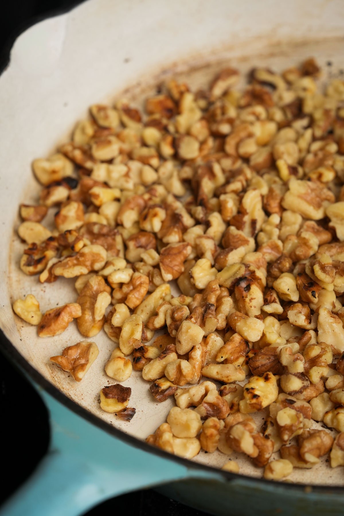 Chopped walnuts being toasted in a light-colored skillet, with some pieces showing slight browning.