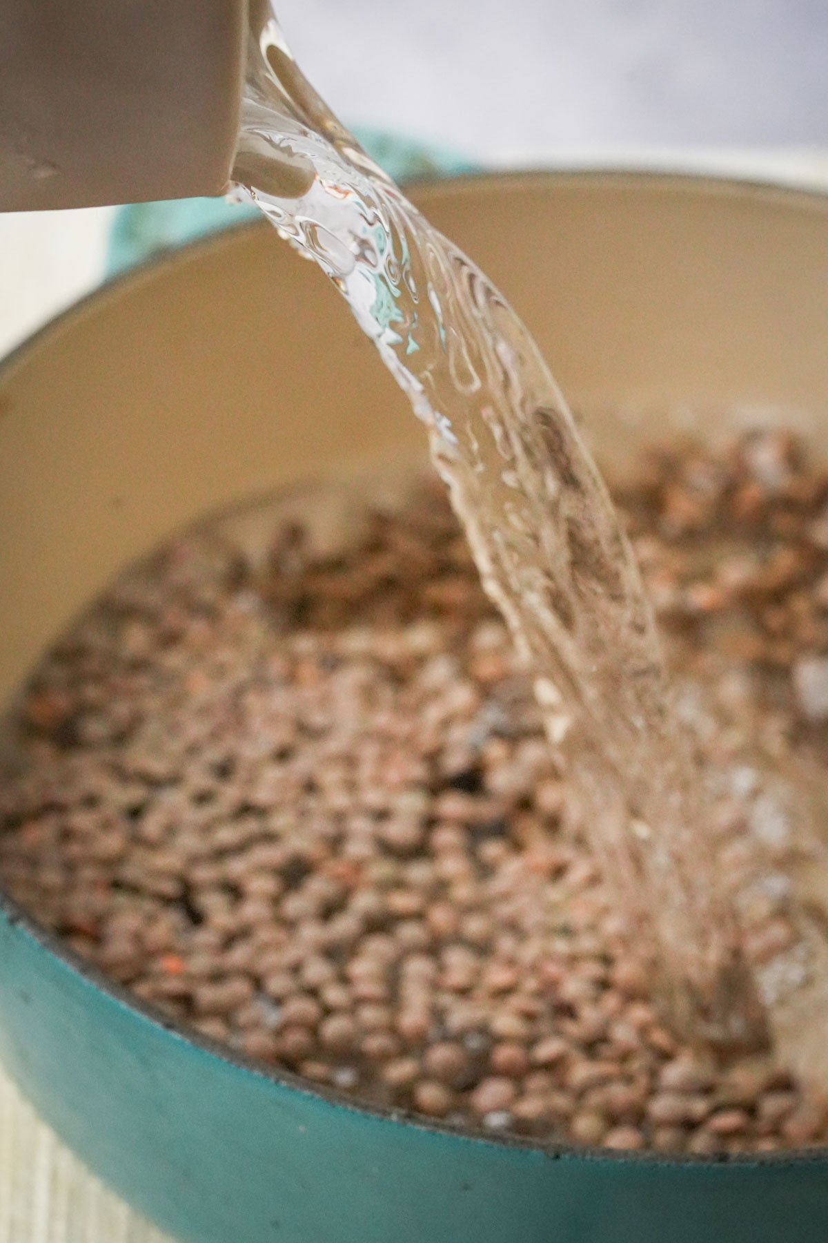 A close-up of water being poured from a container into a teal pot filled with dry lentils, preparing them for cooking.