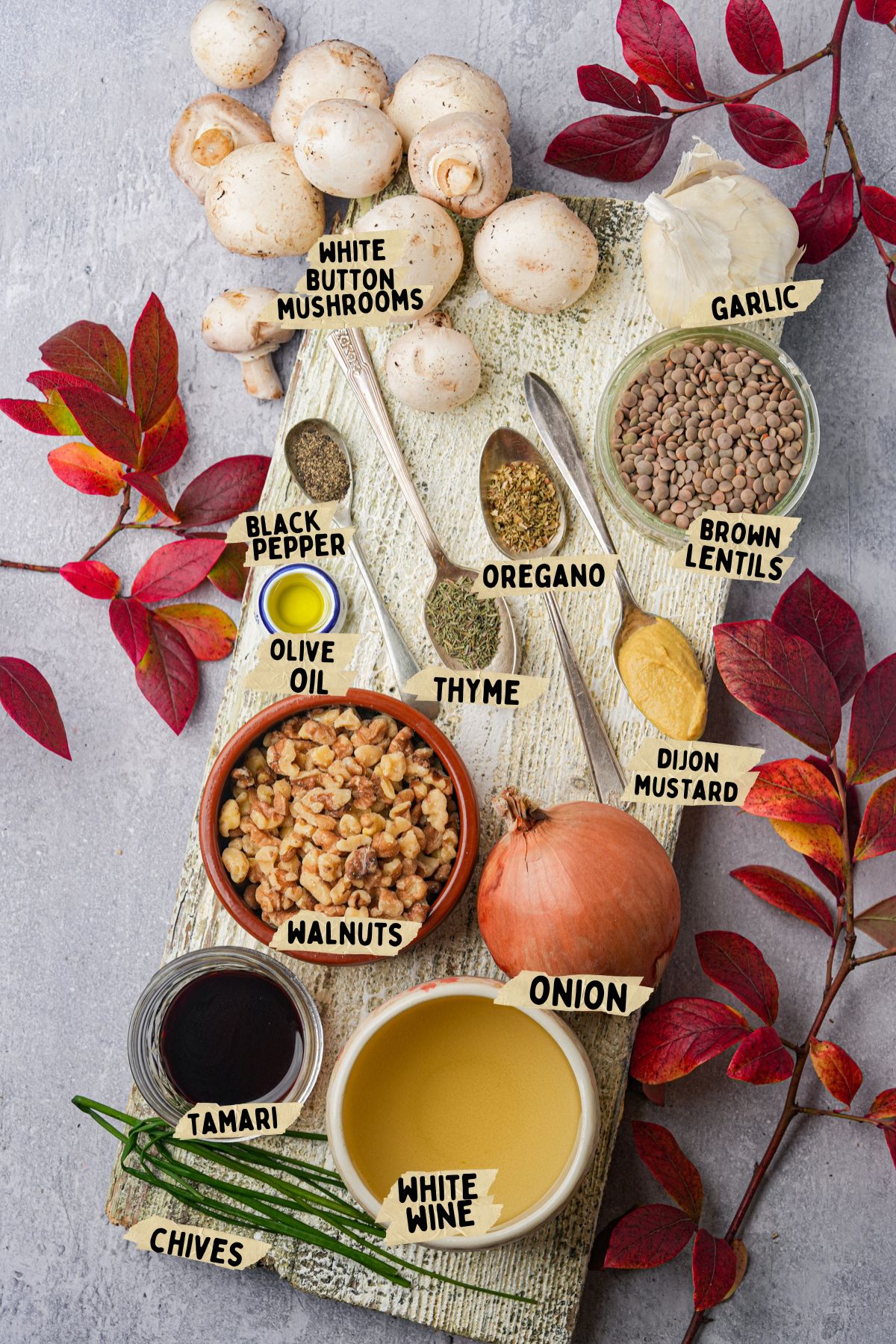 Ingredients for a vegan chopped liver recipe on a board and table, labeled: white button mushrooms, black pepper, olive oil, oregano, thyme, walnuts, tamari, chives, garlic, brown lentils, Dijon mustard, onion, and white wine. Red leaves decorate the scene.