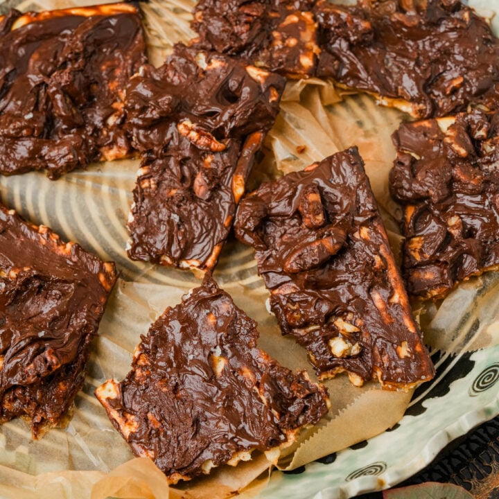 Sliced pieces of chocolate-covered matzo toffee topped with nuts are arranged on parchment paper atop a decorative plate.