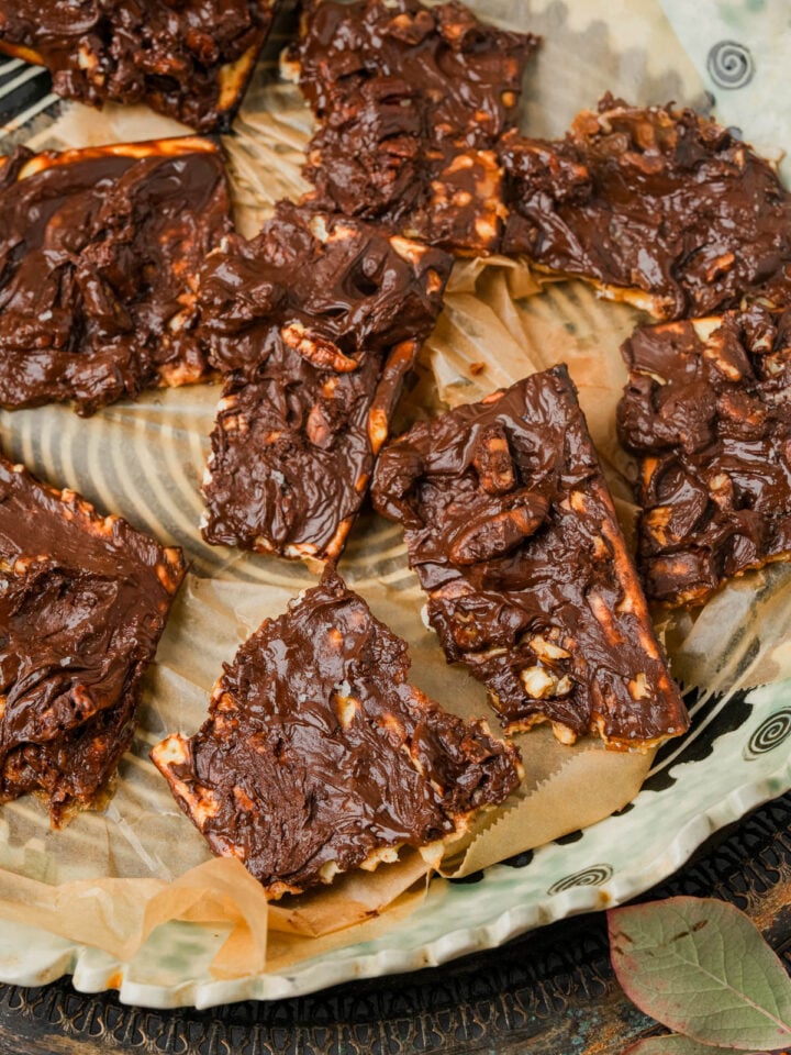 Sliced pieces of chocolate-covered matzo toffee topped with nuts are arranged on parchment paper atop a decorative plate.