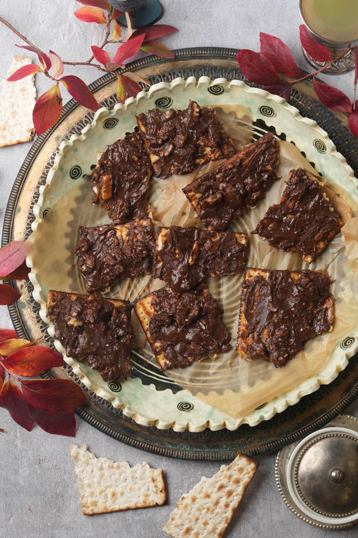 A decorative plate on a silver tray holds pieces of chocolate-covered matzo toffee. Red autumn leaves surround the plate, with broken matzo pieces and a covered dish nearby on a light gray surface.