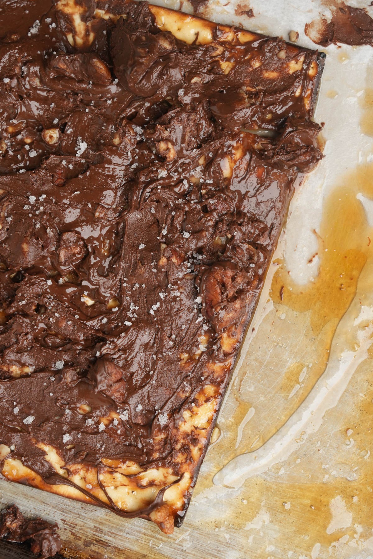 A close-up of a rectangular chocolate-covered vegan matzo toffee on parchment paper, with a rough, textured chocolate layer, visible salt flakes, and some spilled caramel sauce on the side.