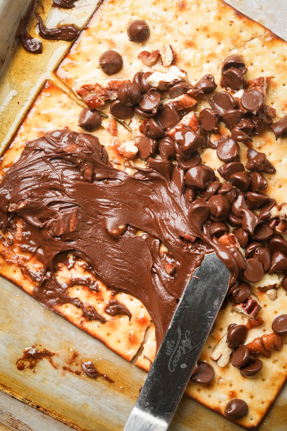 A knife spreads melted chocolate chips and pecans over matzo on a baking sheet.