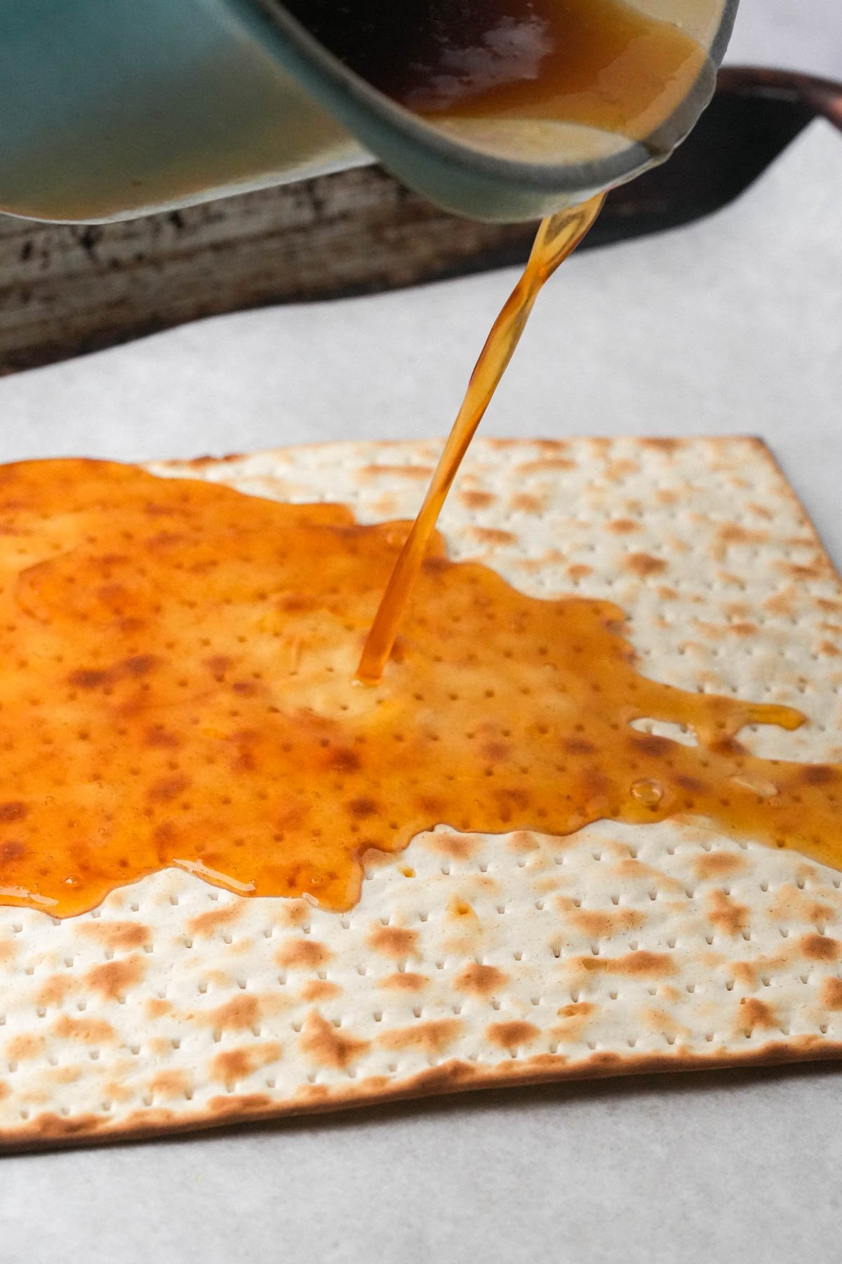 A golden toffee liquid is being poured from a pot onto a large rectangular piece of matzo on a parchment-lined baking sheet.
