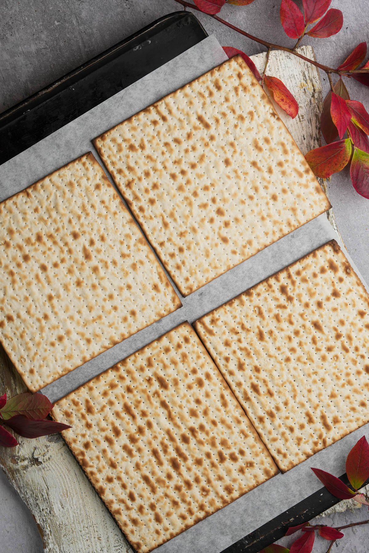 Four square pieces of matzo on a baking sheet lined with parchment paper, surrounded by red autumn leaves on a light textured surface.