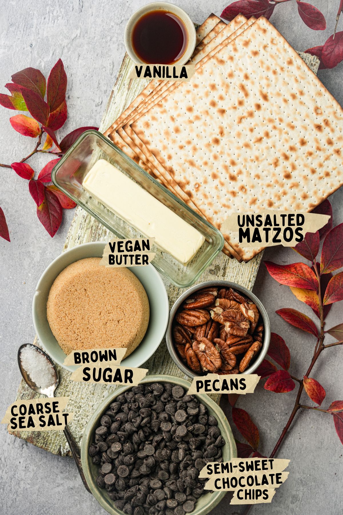 Ingredients for a vegan matzo toffee recipe on a gray background: a stack of unsalted matzos, a bowl of pecans, vegan butter, brown sugar, coarse sea salt, semi-sweet chocolate chips, and a small bowl of vanilla with autumn leaves.