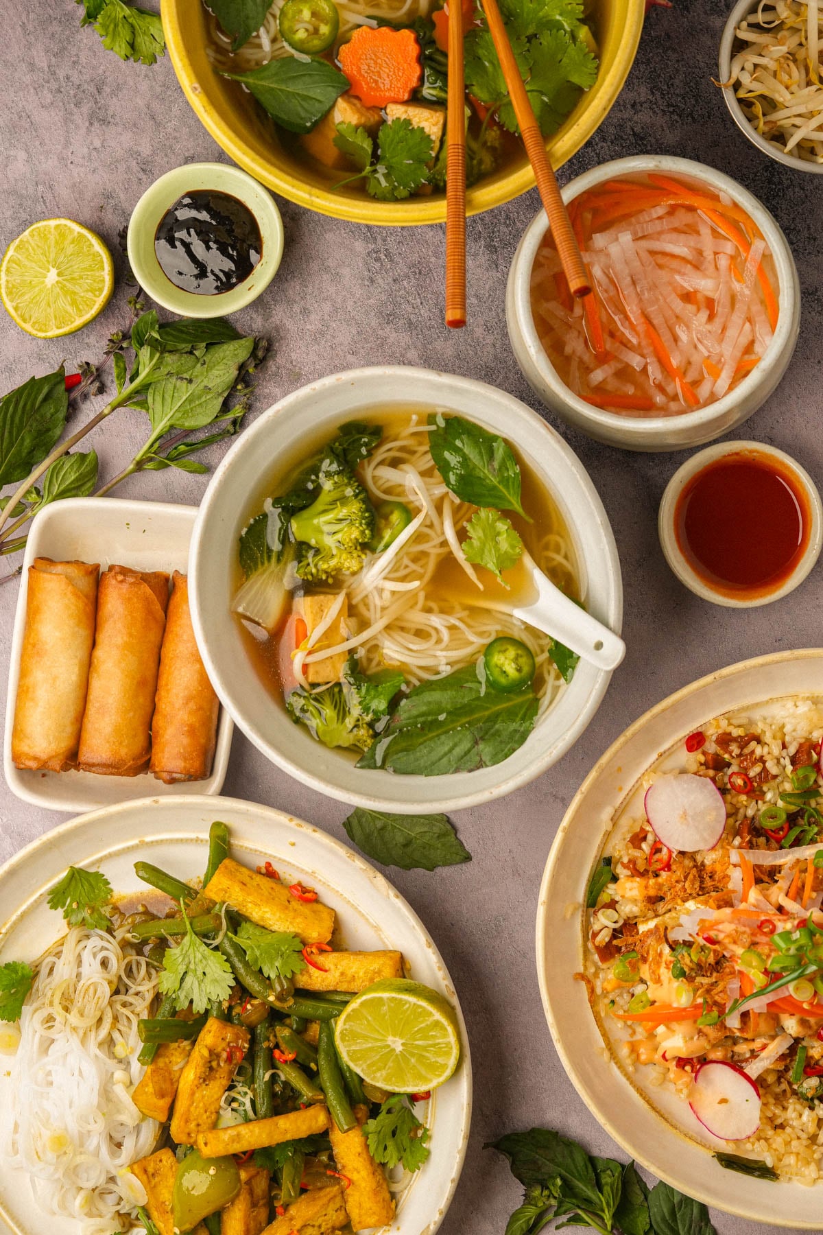 A table of Vietnamese dishes featuring vermicelli noodles with lemongrass tofu and vegetables, spring rolls, vegan pho soup with herbs, Vietnamese sticky rice, fresh lime, dipping sauces, and garnishes on a gray surface.
