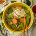 A bowl of vegan pho with broth, noodles, bean sprouts, basil, cilantro, sliced jalapeño, carrot slices, and chopsticks, surrounded by herbs, sauces, and garnishes on a rustic wooden table.