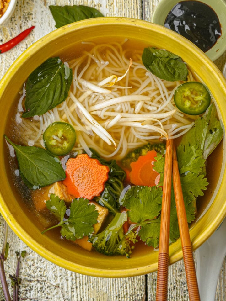 A bowl of vegan pho with broth, noodles, bean sprouts, basil, cilantro, sliced jalapeño, carrot slices, and chopsticks, surrounded by herbs, sauces, and garnishes on a rustic wooden table.