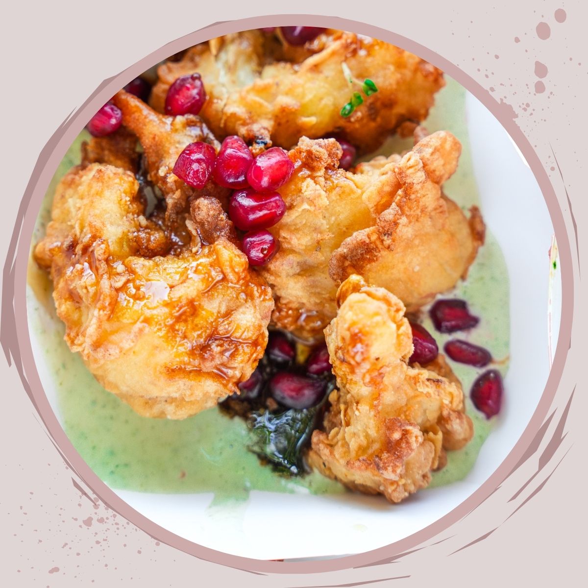 Golden fried cauliflower florets are topped with pomegranate seeds and served on a creamy green sauce, making this Veganuary-friendly dish a vibrant choice, presented in a white bowl with a decorative circular border.