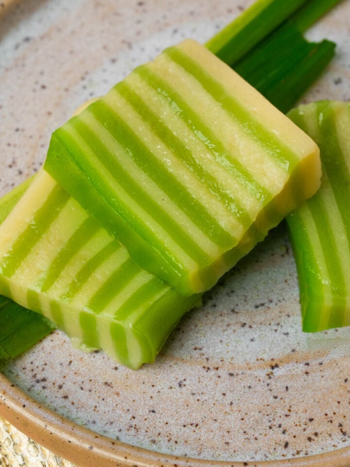 Three pieces of green and cream layered banh da lon dessert are arranged on a speckled ceramic plate with green leaves underneath. The dessert has a striped pattern and glossy texture.