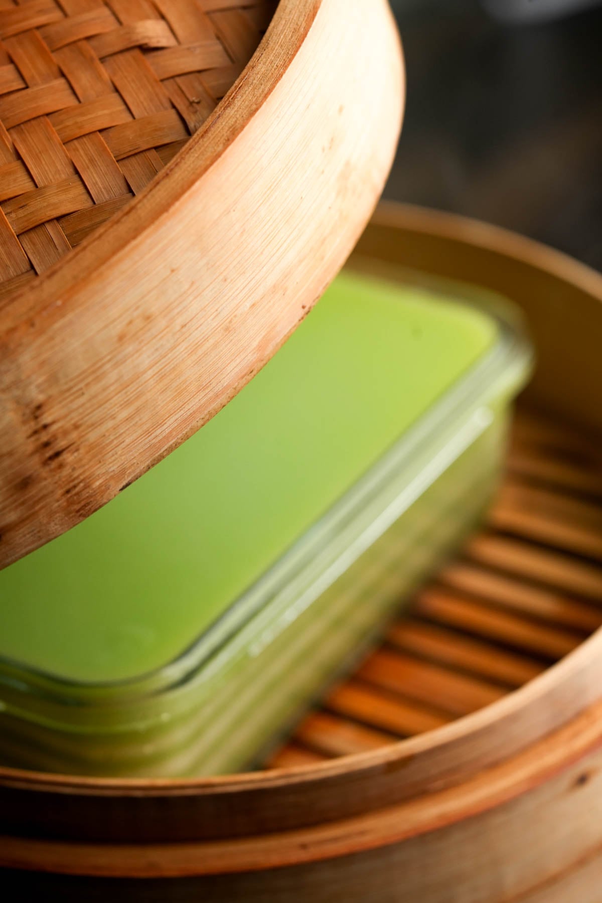 A close-up of a bamboo steamer with the lid partially open, revealing a glass container filled with steamed banh da lon inside, placed on the wooden slats of the steamer basket.