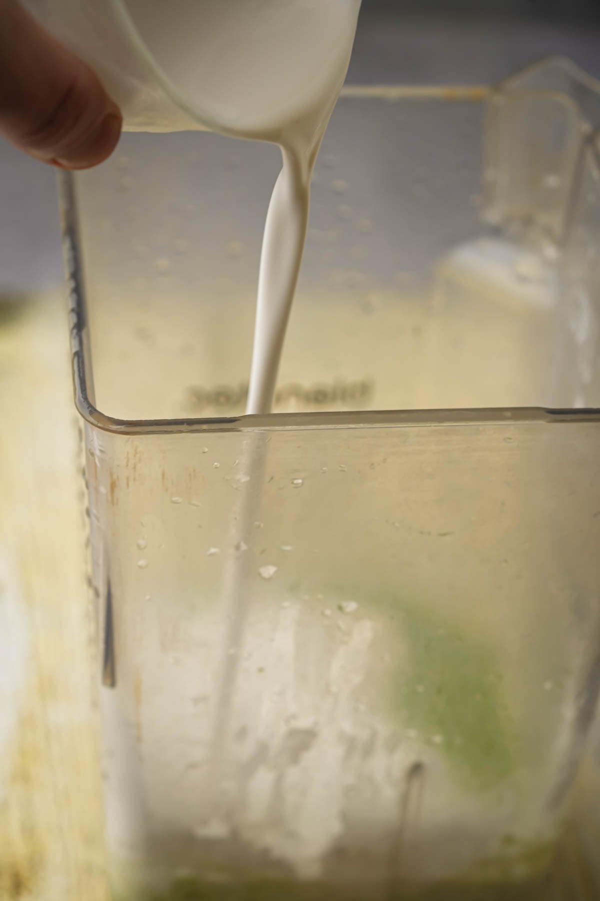 A close-up of coconut milk being poured from a cup into a blender containing a green mixture at the bottom.