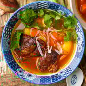 A bowl of Vietnamese vegan bo kho stew with tender mushroom chunks, carrots, potatoes, fresh herbs, chili slices, and onions in a rich, red broth, served in a blue patterned bowl on a wooden table.