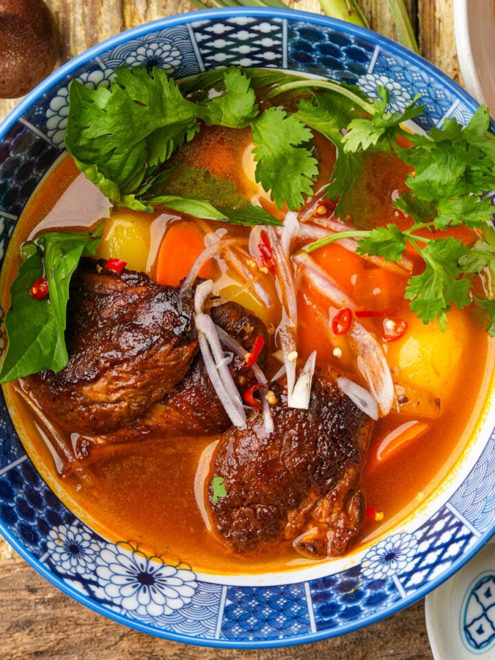 A bowl of Vietnamese vegan bo kho stew with tender mushroom chunks, carrots, potatoes, fresh herbs, chili slices, and onions in a rich, red broth, served in a blue patterned bowl on a wooden table.