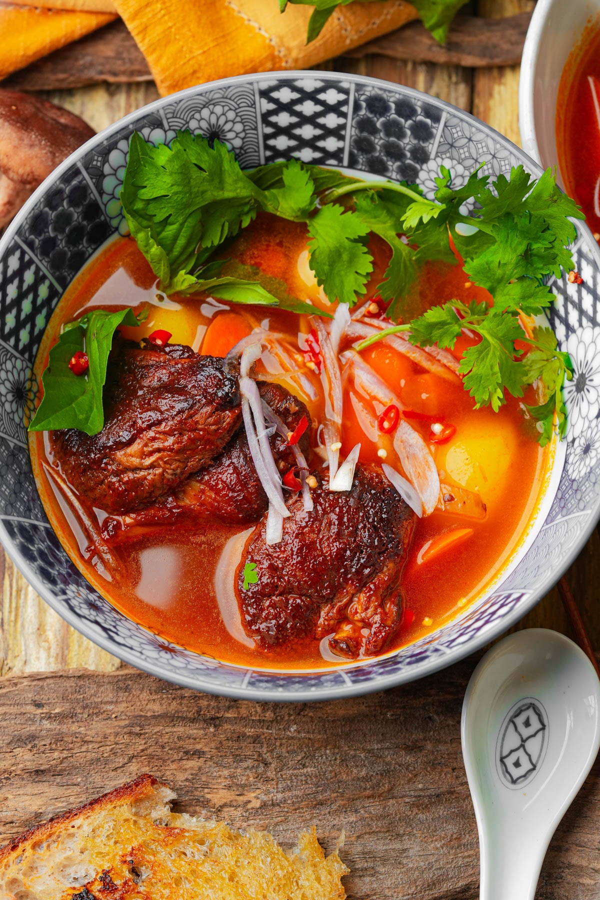 A bowl of Vietnamese vegan bo kho stew with red broth, two pieces of braised mushroom, fresh cilantro, sliced onions, chilies, and herbs, placed on a wooden table beside a spoon and toasted bread.