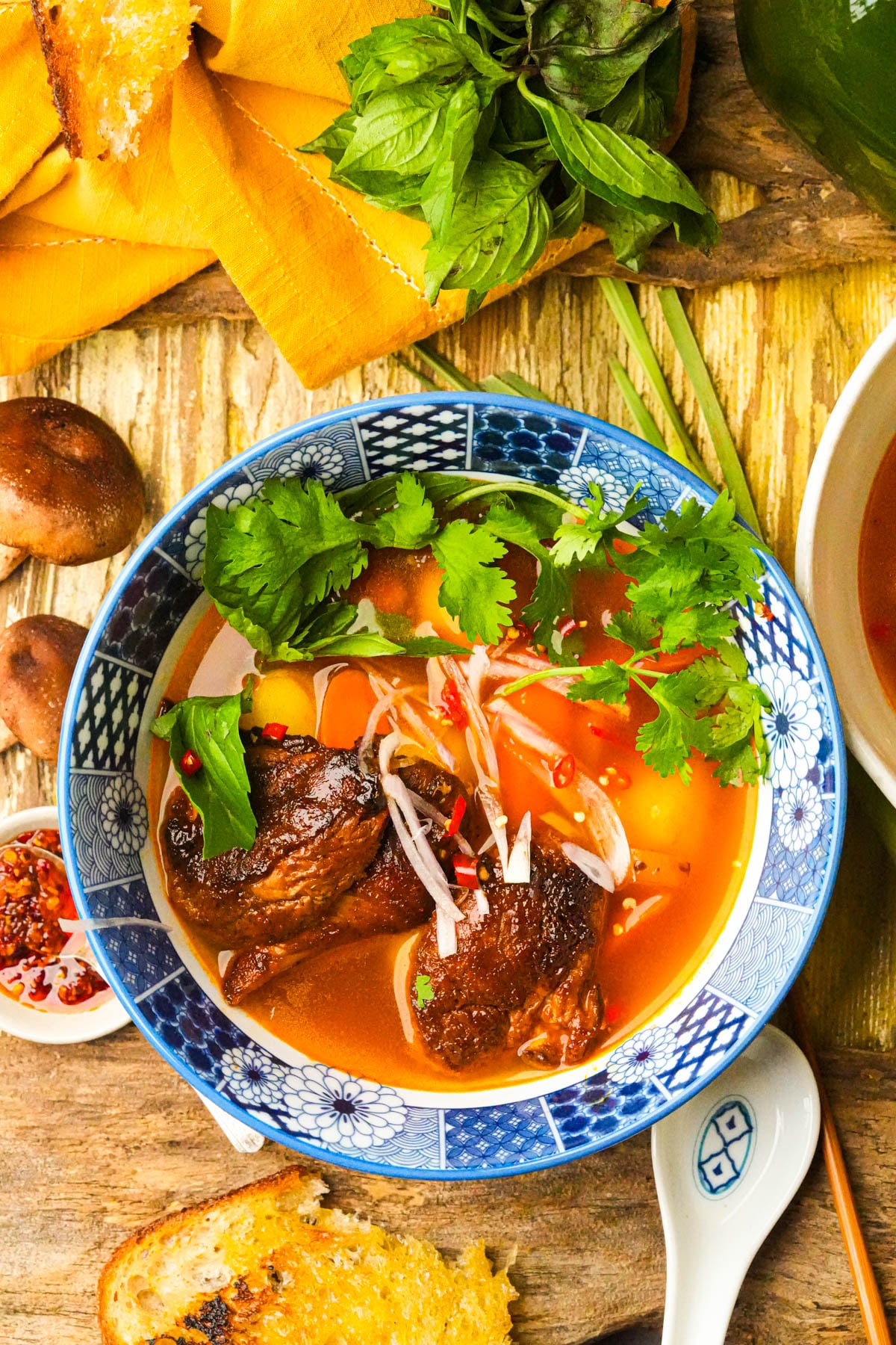 A vibrant bowl of vegan Vietnamese beef stew with tender mushrooms, fresh herbs, and vegetables in a rich red broth, garnished with cilantro. Toasted bread, mushrooms, and leafy greens are arranged on the rustic wooden table.