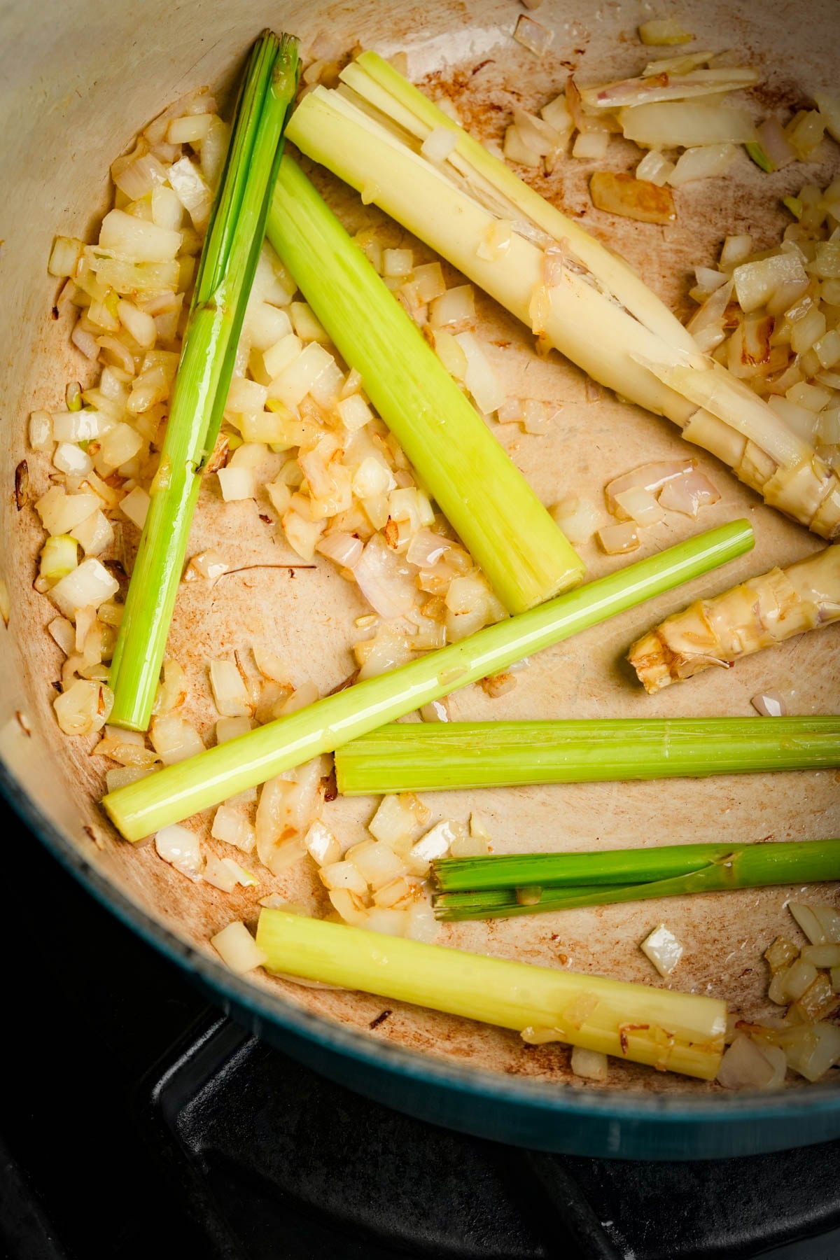 Chopped shallots and stalks of lemongrass are being sautéed in a large pot, with light browning visible on the onions and the pans surface. The pot is set on a stovetop burner.