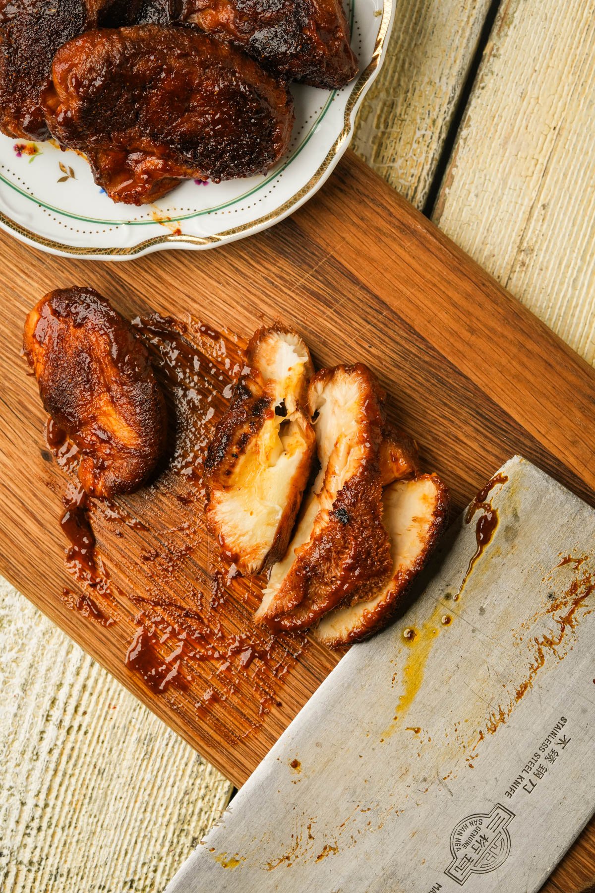 Sliced, glazed cooked mushrooms on a wooden cutting board with a large knife, next to a plate holding more pieces of chicken, all on a rustic light-colored wooden surface.
