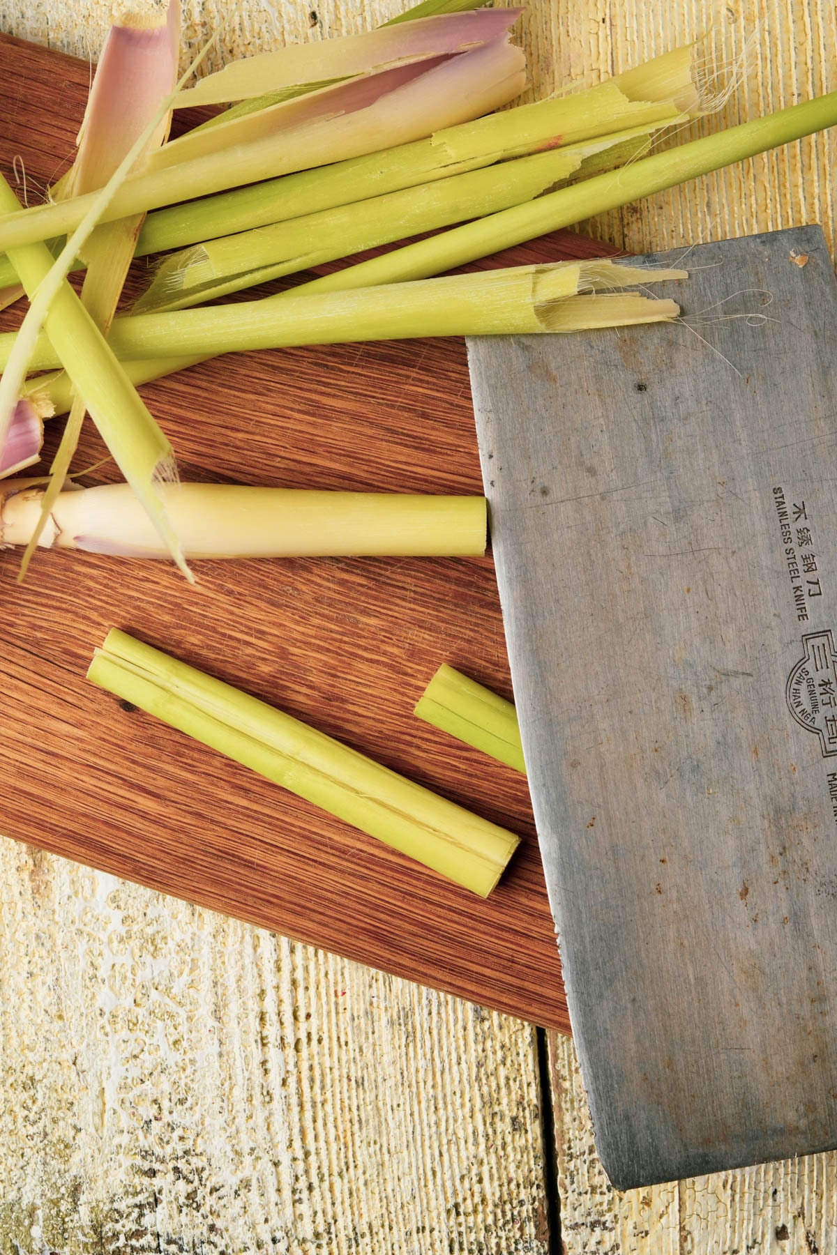 Chopped lemongrass stalks on a wooden cutting board next to a large metal cleaver, on a rustic yellow and white wooden surface.