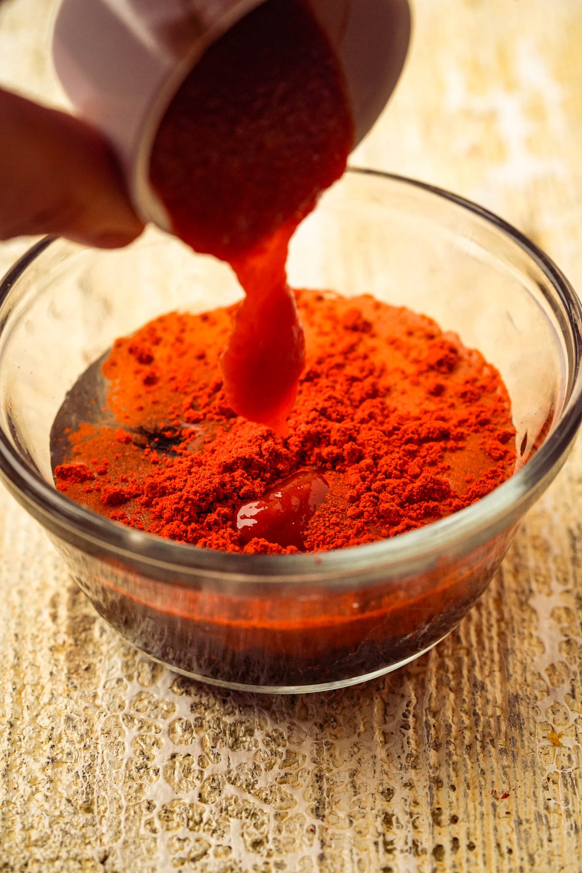 A hand pours sriracha from a small cup into a glass bowl filled with bright red powdered annatto on a light textured surface.