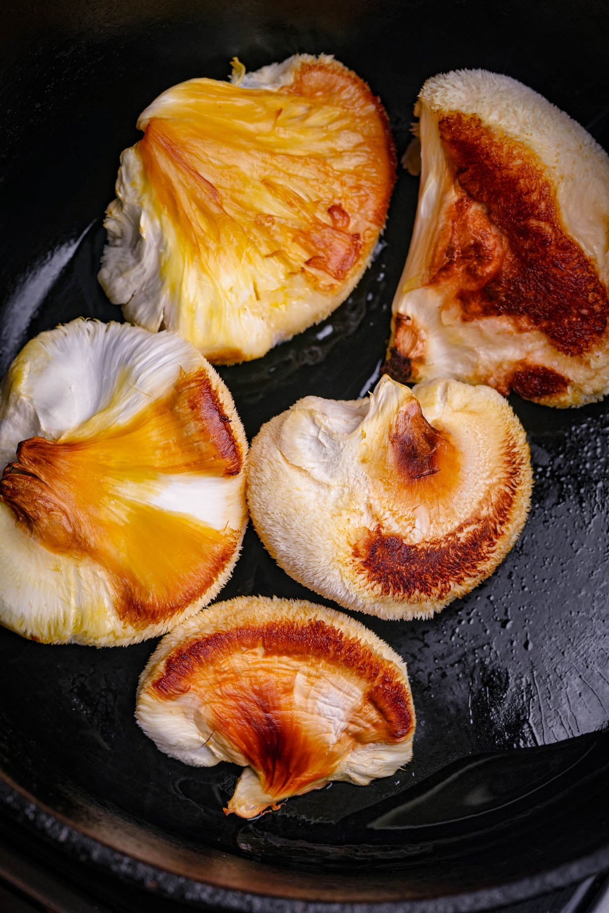 Golden-brown lions mane mushroom pieces sautéing in a pan, edges crisp and slightly charred, with a soft, slightly fuzzy texture and vibrant orange-yellow centers.