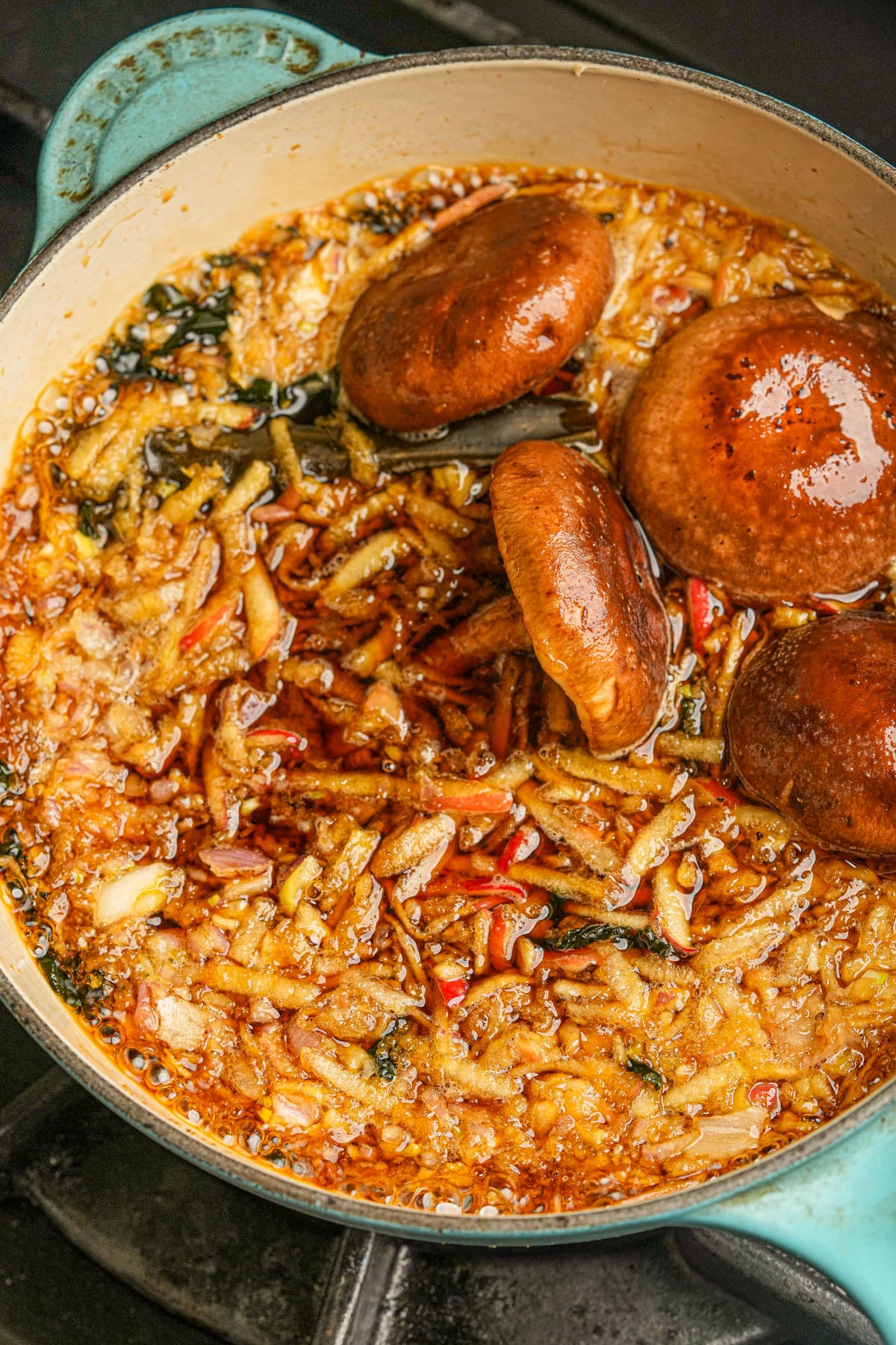 A pot of bubbling Vegan fish sauce with grated apples, sea vegetables, and three whole mushrooms on top, cooking in a rich, golden-brown broth on a stovetop.