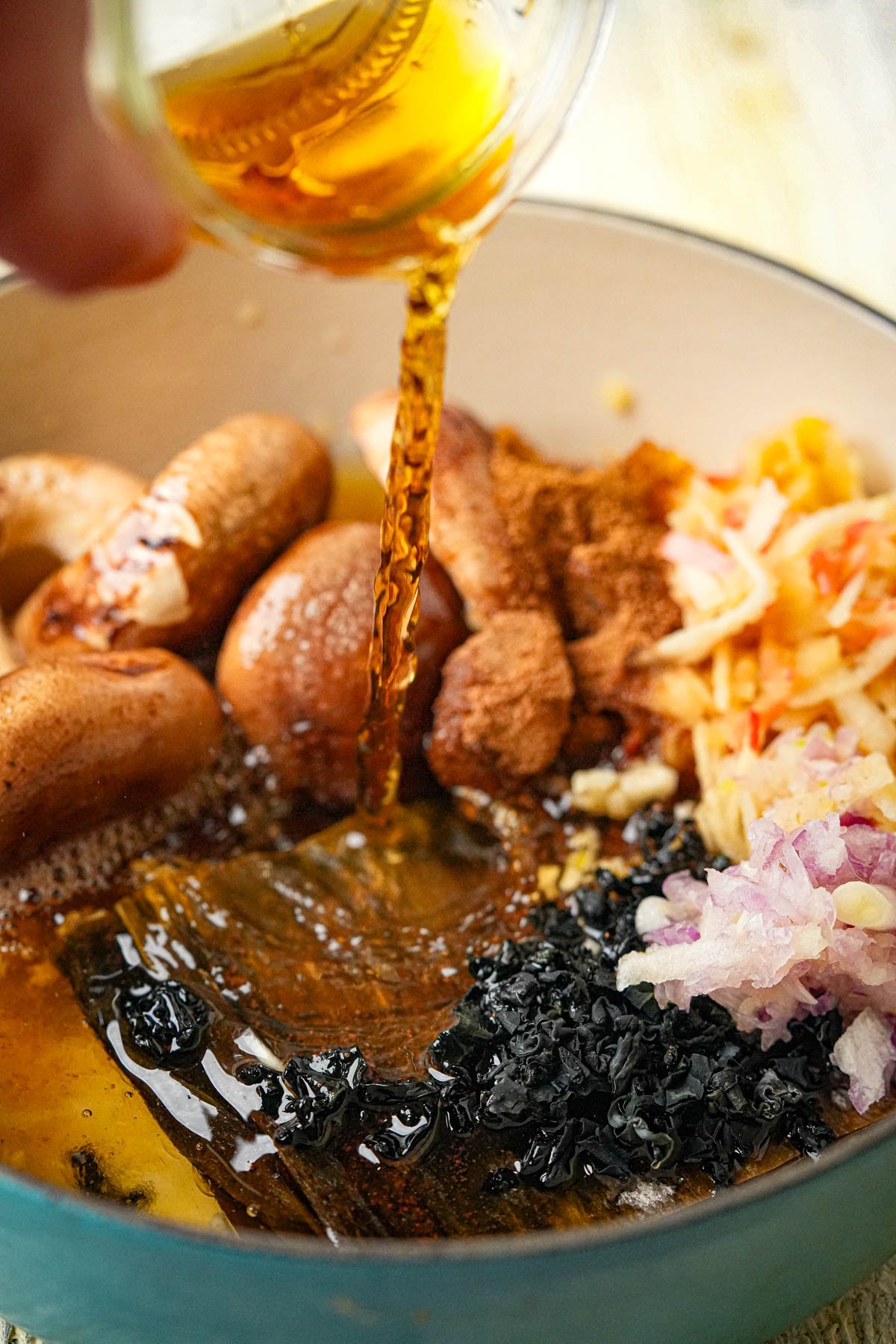 A pitcher of mirin being poured into a pot containing mushrooms, spices, shredded vegetables, chopped onions, and dark seaweed or mushrooms, ready to be cooked together to form a batch of Vegan fish sauce.