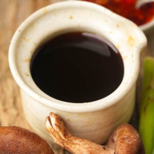 A small ceramic pitcher filled with Vegan fish sauce surrounded by fresh mushrooms and green vegetables on a wooden surface.
