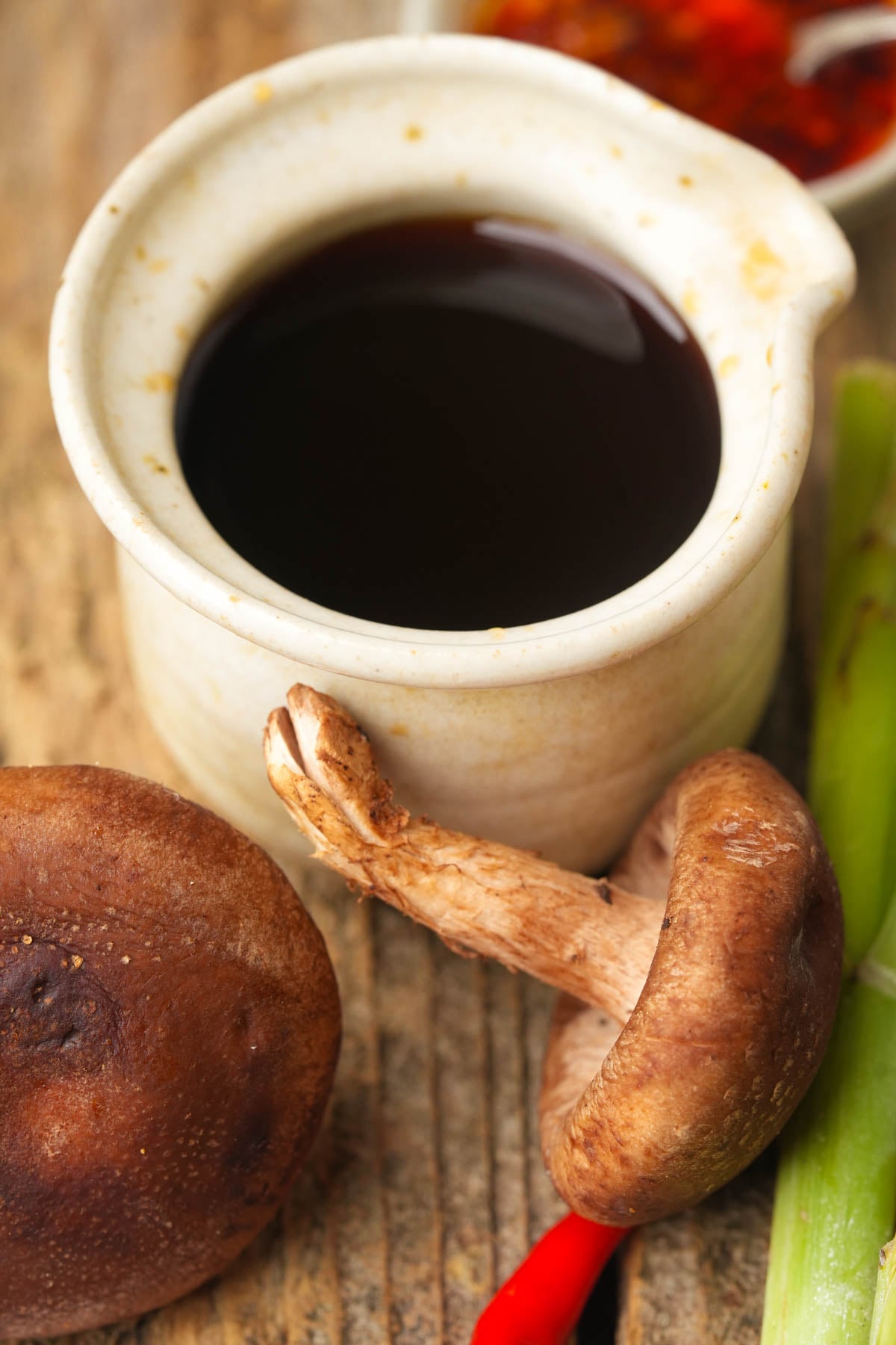 A small ceramic cup filled with Vegan fish sauce surrounded by shiitake mushrooms, a red chili pepper, a green onion, and a bowl of sauce, all on a rustic wooden surface.
