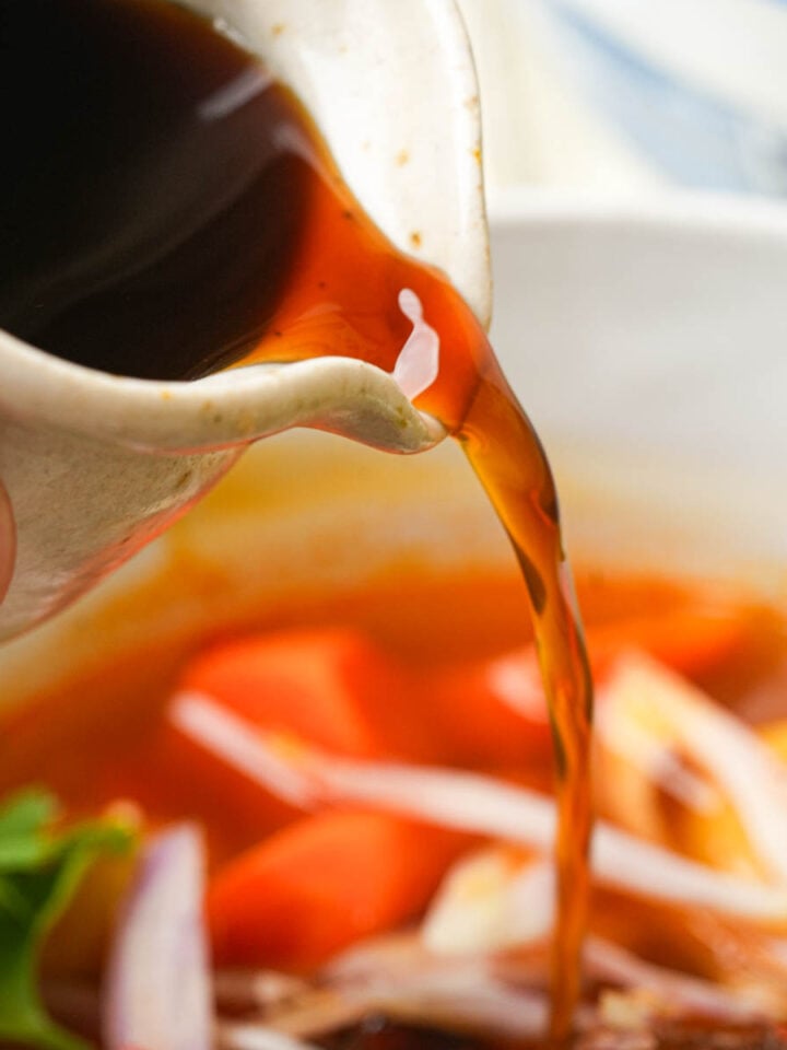 Vegan fish sauce being poured from a small ceramic cup into a bowl of soup with visible vegetables and herbs.