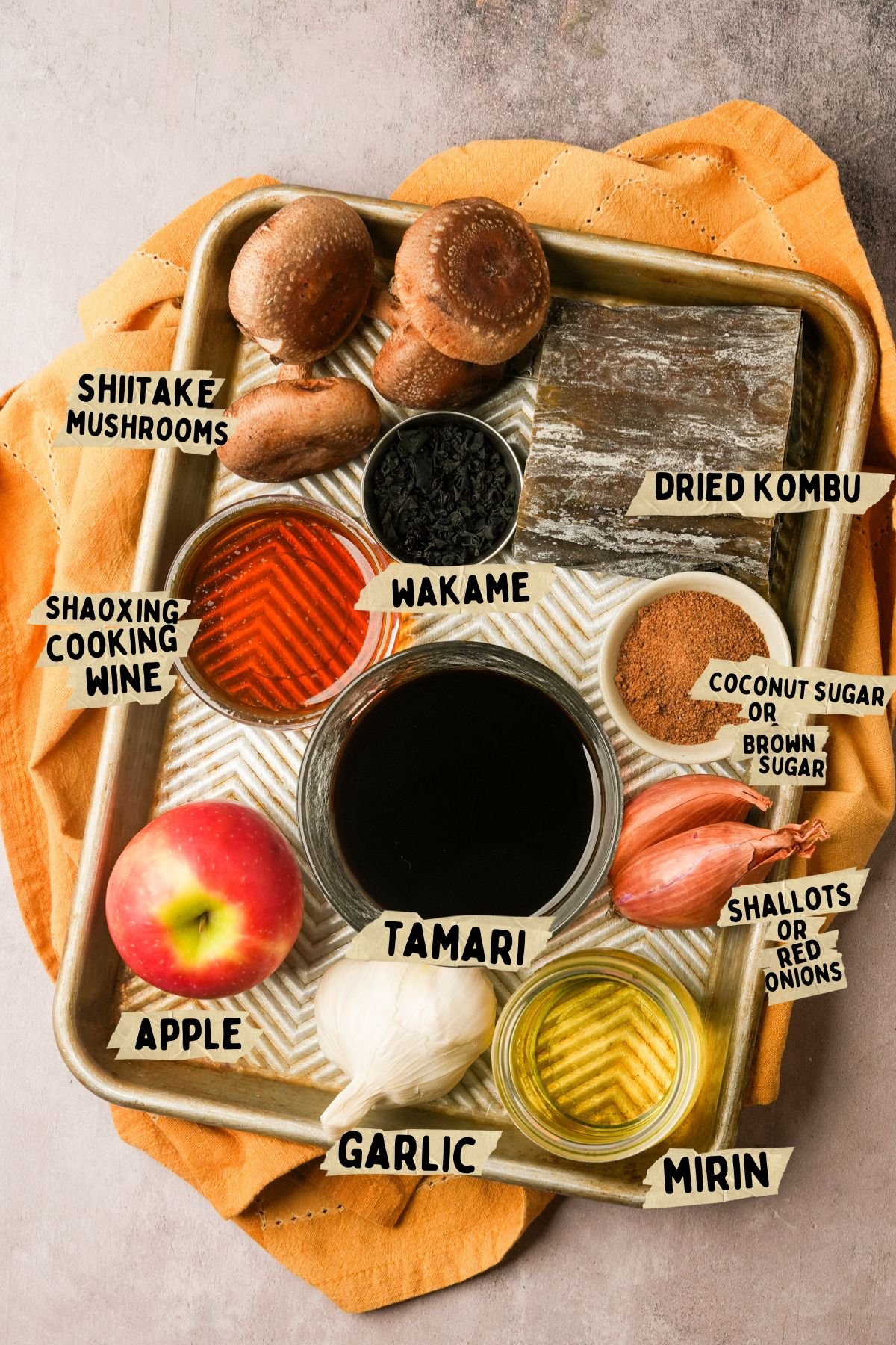 A metal tray with labeled ingredients for a Vegan fish sauce recipe: shiitake mushrooms, dried kombu, wakame, Shaoxing cooking wine, coconut sugar, shallots, tamari, apple, garlic, and mirin, arranged on an orange cloth.