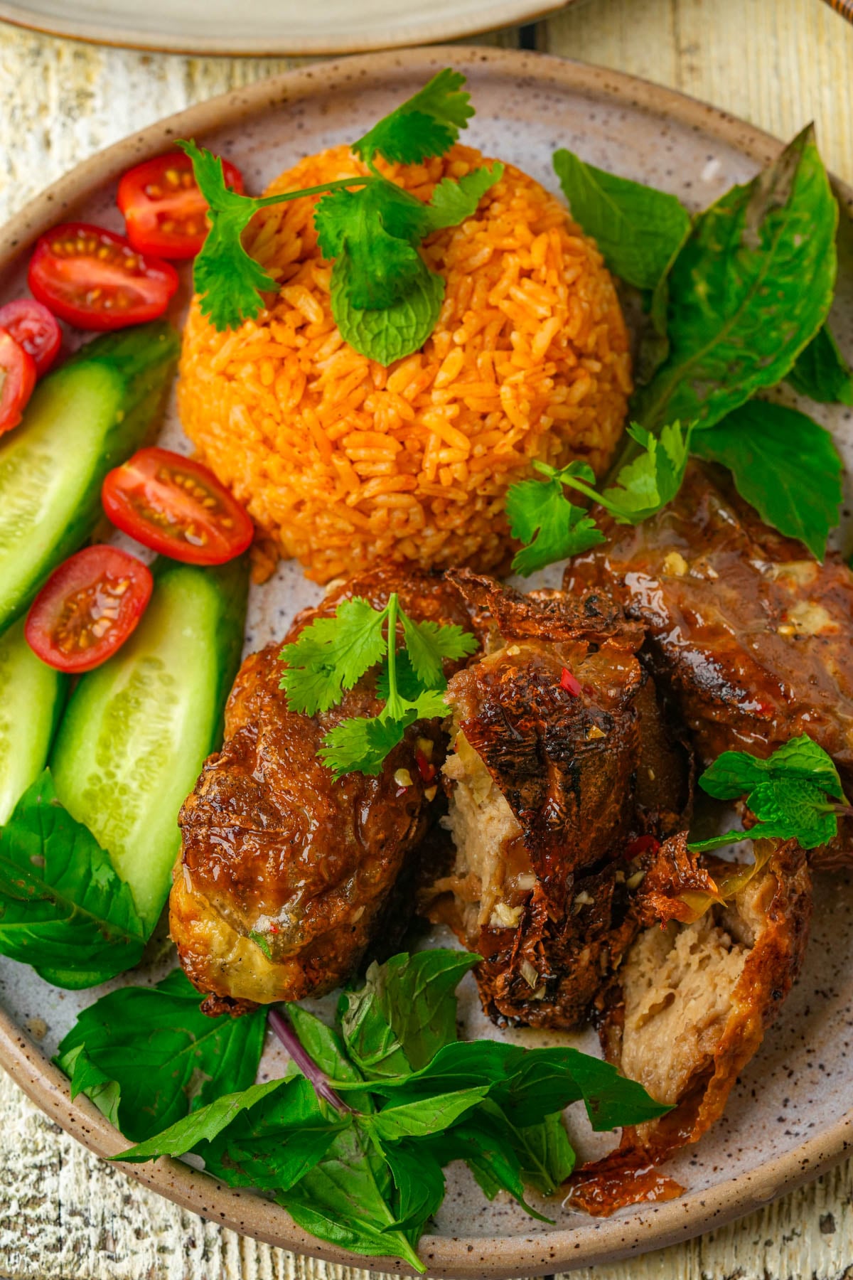 A plate with a mound of orange rice, vegan Vietnamese fried chicken, cucumber sticks, halved cherry tomatoes, and fresh herbs, including cilantro and basil, arranged on a speckled ceramic dish.