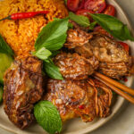 A plate of vegan Vietnamese fried chicken with herbs, red rice, fresh lime, cherry tomatoes, and garnished with basil and mint leaves, with chopsticks resting on the side.