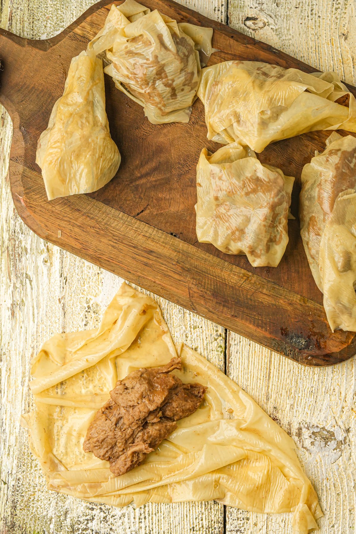 Six pieces of yuba with a seitan filling are arranged on a rustic wooden board, with one open piece of yuba and filling on a white, weathered wooden surface below for forming vegan Vietnamese fried chicken.