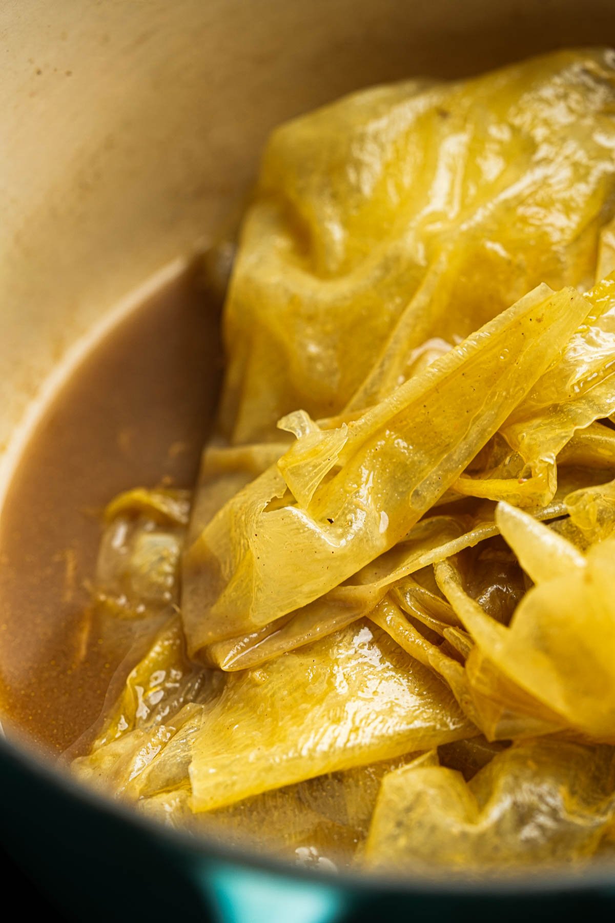 Close-up of yuba sheets in a brown broth inside a pot. The yuba appears soft and tender with visible folds in the sheets.
