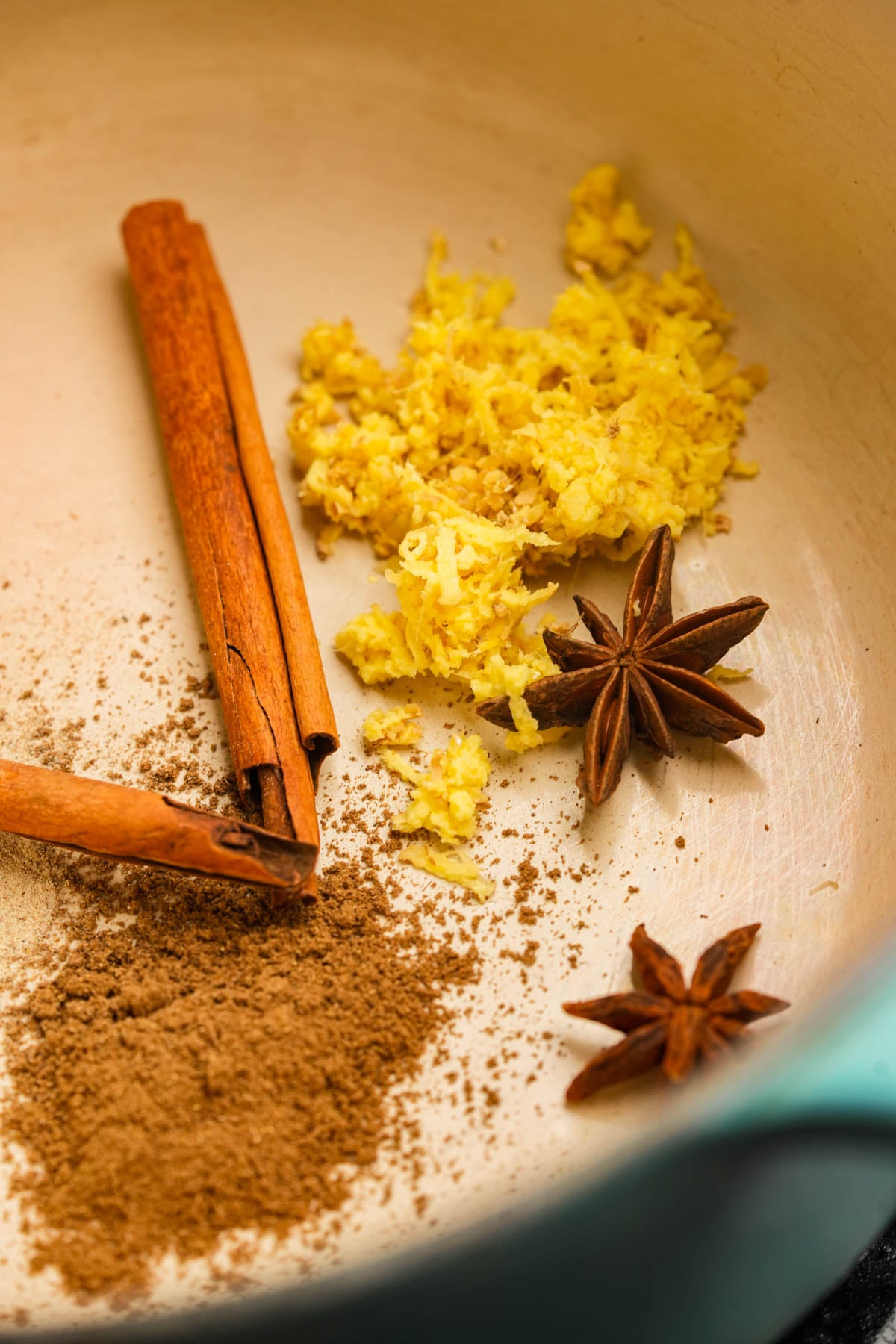 A close-up of a pot containing ground spices, grated ginger, two cinnamon sticks, and two star anise pods arranged on the surface.