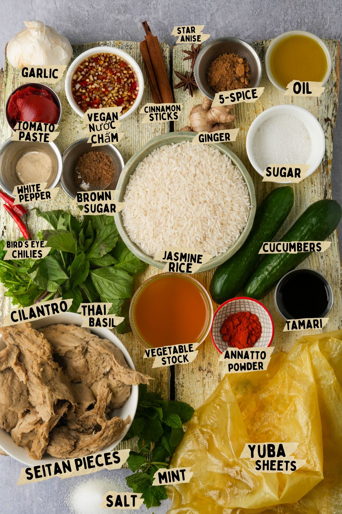 A variety of labeled vegan ingredients for a vegan Vietnamese fried chicken recipe are arranged on a rustic surface, including garlic, spices, jasmine rice, cucumbers, seitan, yuba sheets, herbs, sauces, sugars, and oil.