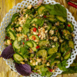 A bowl of sliced cucumber salad topped with chopped peanuts, red chili slices, fresh herbs, and leafy greens, served in a white patterned bowl on a rustic wooden surface.