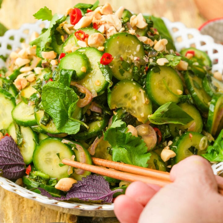 A hand holding chopsticks reaches for a fresh cucumber salad garnished with chopped peanuts, herbs, red chili slices, and leafy greens, served in a white decorative bowl.